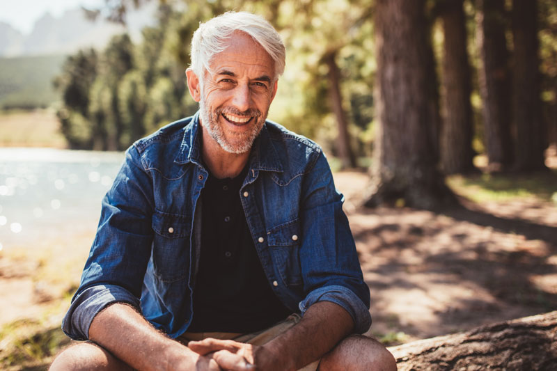Middle age man sitting outdoors near a forest, smiling at camera